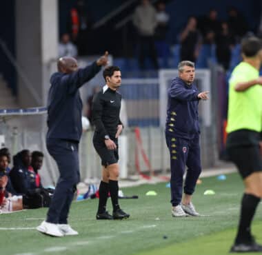 Football sideline scene: coach in black with headset stands while another coach in a navy tracksuit points, substitutes in red sit on the bench.