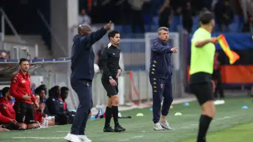 Football sideline scene: coach in black with headset stands while another coach in a navy tracksuit points, substitutes in red sit on the bench.