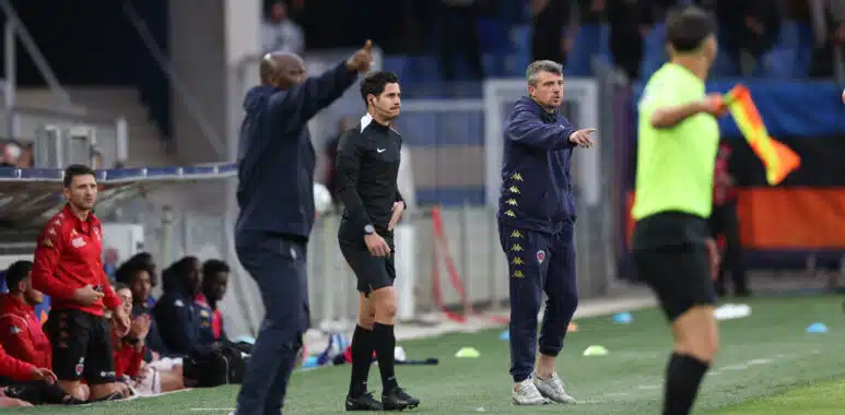 Football sideline scene: coach in black with headset stands while another coach in a navy tracksuit points, substitutes in red sit on the bench.