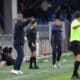 Football sideline scene: coach in black with headset stands while another coach in a navy tracksuit points, substitutes in red sit on the bench.
