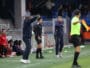 Football sideline scene: coach in black with headset stands while another coach in a navy tracksuit points, substitutes in red sit on the bench.