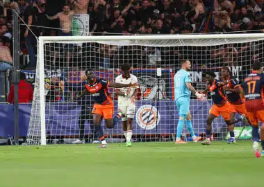 Soccer players in orange and navy celebrate near the goal as fans watch from the stands behind the net.