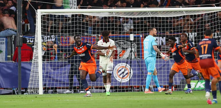 Soccer players in orange and navy celebrate near the goal as fans watch from the stands behind the net.