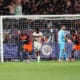Soccer players in orange and navy celebrate near the goal as fans watch from the stands behind the net.