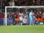 Soccer players in orange and navy celebrate near the goal as fans watch from the stands behind the net.