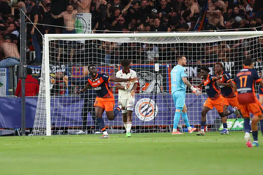 Mendy | AllezPaillade.com Soccer players in orange and navy celebrate near the goal as fans watch from the stands behind the net.