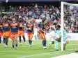 Soccer players in orange and navy kits celebrate near the goal as a goalkeeper looks on from the right with a net behind them and a crowded stadium in the background.