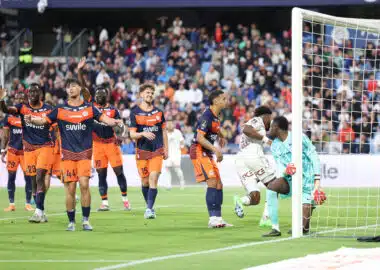 Soccer players in orange and navy kits celebrate near the goal as a goalkeeper looks on from the right with a net behind them and a crowded stadium in the background.