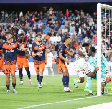Soccer players in orange and navy kits celebrate near the goal as a goalkeeper looks on from the right with a net behind them and a crowded stadium in the background.