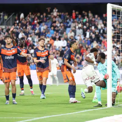 Soccer players in orange and navy kits celebrate near the goal as a goalkeeper looks on from the right with a net behind them and a crowded stadium in the background.