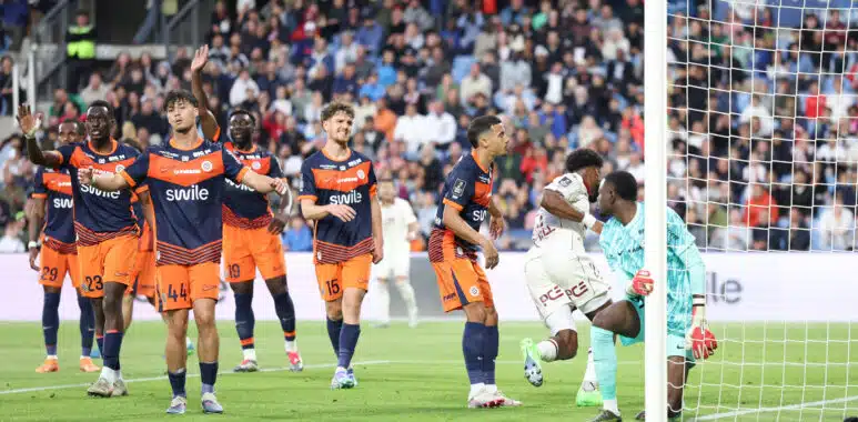 Soccer players in orange and navy kits celebrate near the goal as a goalkeeper looks on from the right with a net behind them and a crowded stadium in the background.