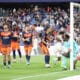 Soccer players in orange and navy kits celebrate near the goal as a goalkeeper looks on from the right with a net behind them and a crowded stadium in the background.