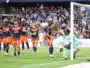 Soccer players in orange and navy kits celebrate near the goal as a goalkeeper looks on from the right with a net behind them and a crowded stadium in the background.