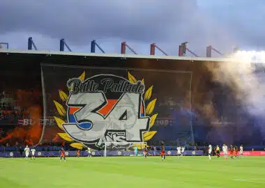 Large stadium banner reading 'Butte Paillade 34' with yellow laurel leaves behind a soccer goal; players on the pitch and orange smoke in the stands.