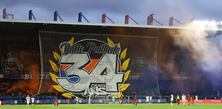 Large stadium banner reading 'Butte Paillade 34' with yellow laurel leaves behind a soccer goal; players on the pitch and orange smoke in the stands.