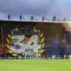 Large stadium banner reading 'Butte Paillade 34' with yellow laurel leaves behind a soccer goal; players on the pitch and orange smoke in the stands.