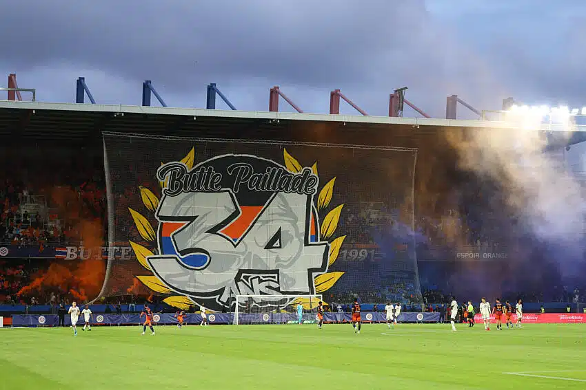 Large stadium banner reading 'Butte Paillade 34' with yellow laurel leaves behind a soccer goal; players on the pitch and orange smoke in the stands.