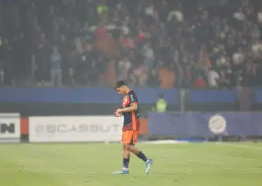 Soccer player in orange and navy kit walking on a grass field with a blurred stadium crowd in the background, looking down at the ground.