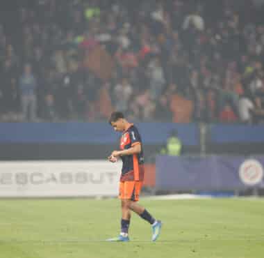 Soccer player in orange and navy kit walking on a grass field with a blurred stadium crowd in the background, looking down at the ground.
