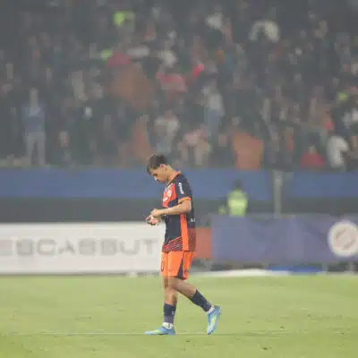 Soccer player in orange and navy kit walking on a grass field with a blurred stadium crowd in the background, looking down at the ground.