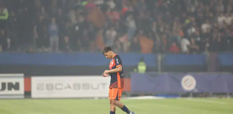 Soccer player in orange and navy kit walking on a grass field with a blurred stadium crowd in the background, looking down at the ground.