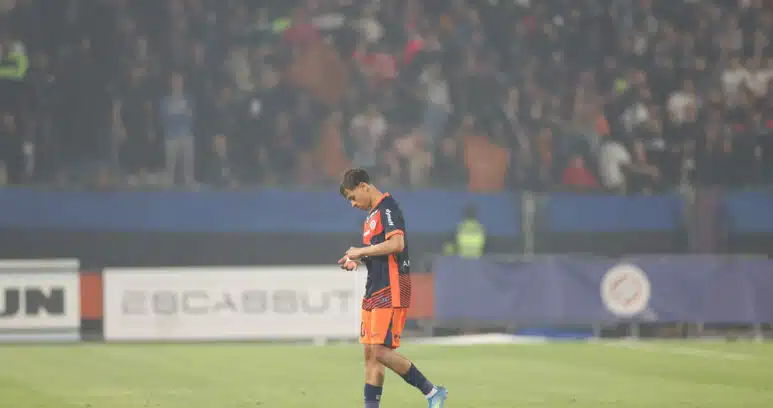 Soccer player in orange and navy kit walking on a grass field with a blurred stadium crowd in the background, looking down at the ground.