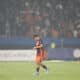Soccer player in orange and navy kit walking on a grass field with a blurred stadium crowd in the background, looking down at the ground.