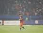 Soccer player in orange and navy kit walking on a grass field with a blurred stadium crowd in the background, looking down at the ground.