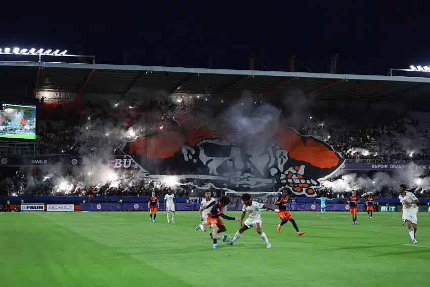 Nighttime soccer match with players on a lit green field and a huge orange-and-black banner featuring a skull in the stands, smoke flares rising from the crowd.