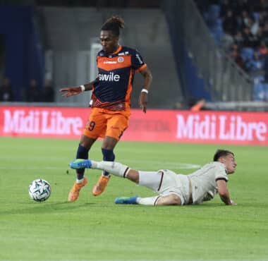 Soccer player in an orange and navy kit controls the ball while a white-clad opponent slides on the field in a stadium crowd backdrop.