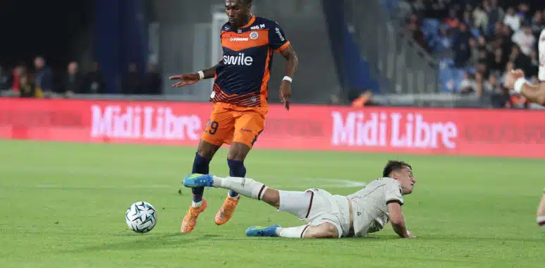 Tchato Montpellier Herault SC v Clermont Foot 63 - Ligue 2 BKT 20252026 | AllezPaillade.com Soccer player in an orange and navy kit controls the ball while a white-clad opponent slides on the field in a stadium crowd backdrop.