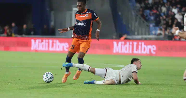 Soccer player in an orange and navy kit controls the ball while a white-clad opponent slides on the field in a stadium crowd backdrop.