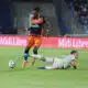 Soccer player in an orange and navy kit controls the ball while a white-clad opponent slides on the field in a stadium crowd backdrop.