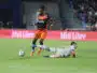 Soccer player in an orange and navy kit controls the ball while a white-clad opponent slides on the field in a stadium crowd backdrop.