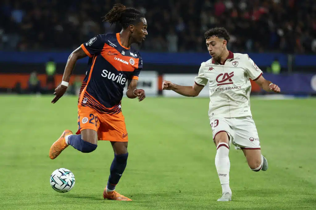 Two soccer players race for the ball on a green pitch during a match, one wearing navy and orange and the other white.