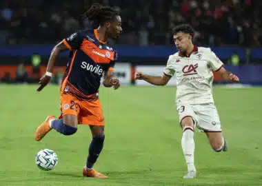 Two soccer players race for the ball on a green pitch during a match, one wearing navy and orange and the other white.