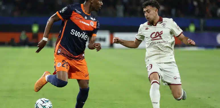 Two soccer players race for the ball on a green pitch during a match, one wearing navy and orange and the other white.