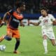 Two soccer players race for the ball on a green pitch during a match, one wearing navy and orange and the other white.
