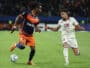 Two soccer players race for the ball on a green pitch during a match, one wearing navy and orange and the other white.