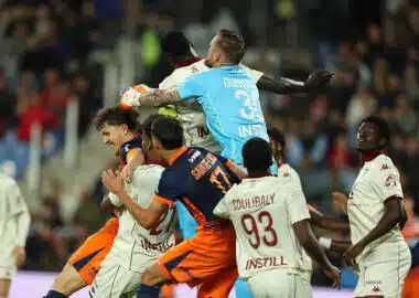 Group of soccer players struggle for the ball in a crowded match, with a goalkeeper in a light-blue kit leaping to punch the ball away.