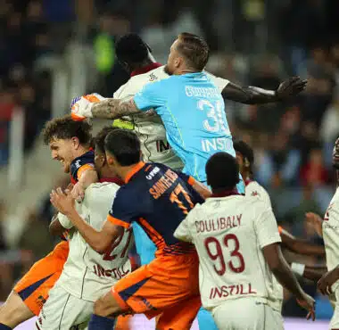 Group of soccer players struggle for the ball in a crowded match, with a goalkeeper in a light-blue kit leaping to punch the ball away.