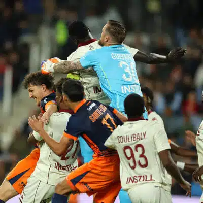 Group of soccer players struggle for the ball in a crowded match, with a goalkeeper in a light-blue kit leaping to punch the ball away.