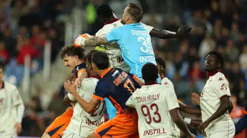Group of soccer players struggle for the ball in a crowded match, with a goalkeeper in a light-blue kit leaping to punch the ball away.