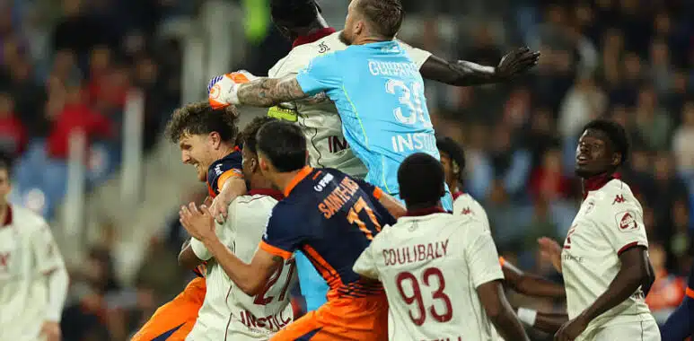 Group of soccer players struggle for the ball in a crowded match, with a goalkeeper in a light-blue kit leaping to punch the ball away.