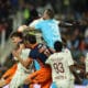 Group of soccer players struggle for the ball in a crowded match, with a goalkeeper in a light-blue kit leaping to punch the ball away.