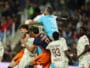 Group of soccer players struggle for the ball in a crowded match, with a goalkeeper in a light-blue kit leaping to punch the ball away.