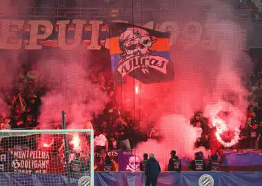Crowd of football fans in a stadium, red smoke from flares, with a banner featuring a skull and the word 'Ultras' above the goal.