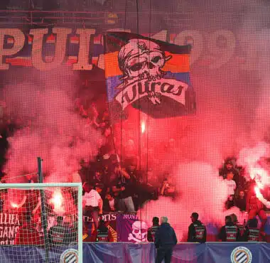 Crowd of football fans in a stadium, red smoke from flares, with a banner featuring a skull and the word 'Ultras' above the goal.