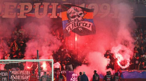 Crowd of football fans in a stadium, red smoke from flares, with a banner featuring a skull and the word 'Ultras' above the goal.