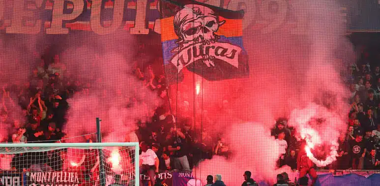 Crowd of football fans in a stadium, red smoke from flares, with a banner featuring a skull and the word 'Ultras' above the goal.